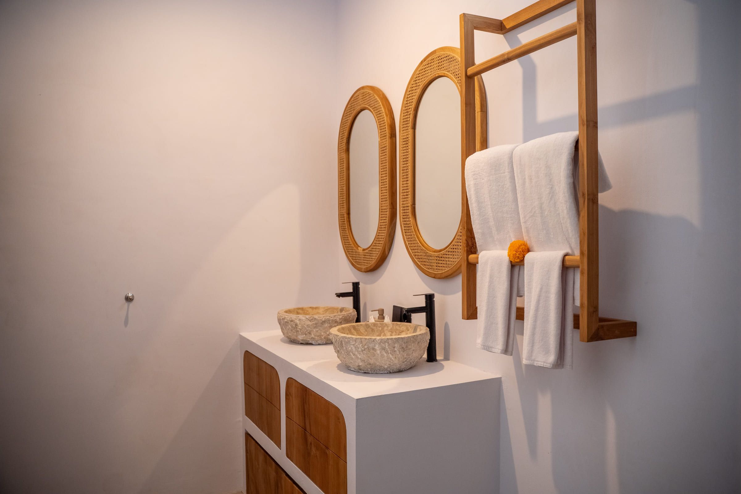 Bathroom vanity with twin stone basins, rattan-framed oval mirrors and folded towels on a wooden rail