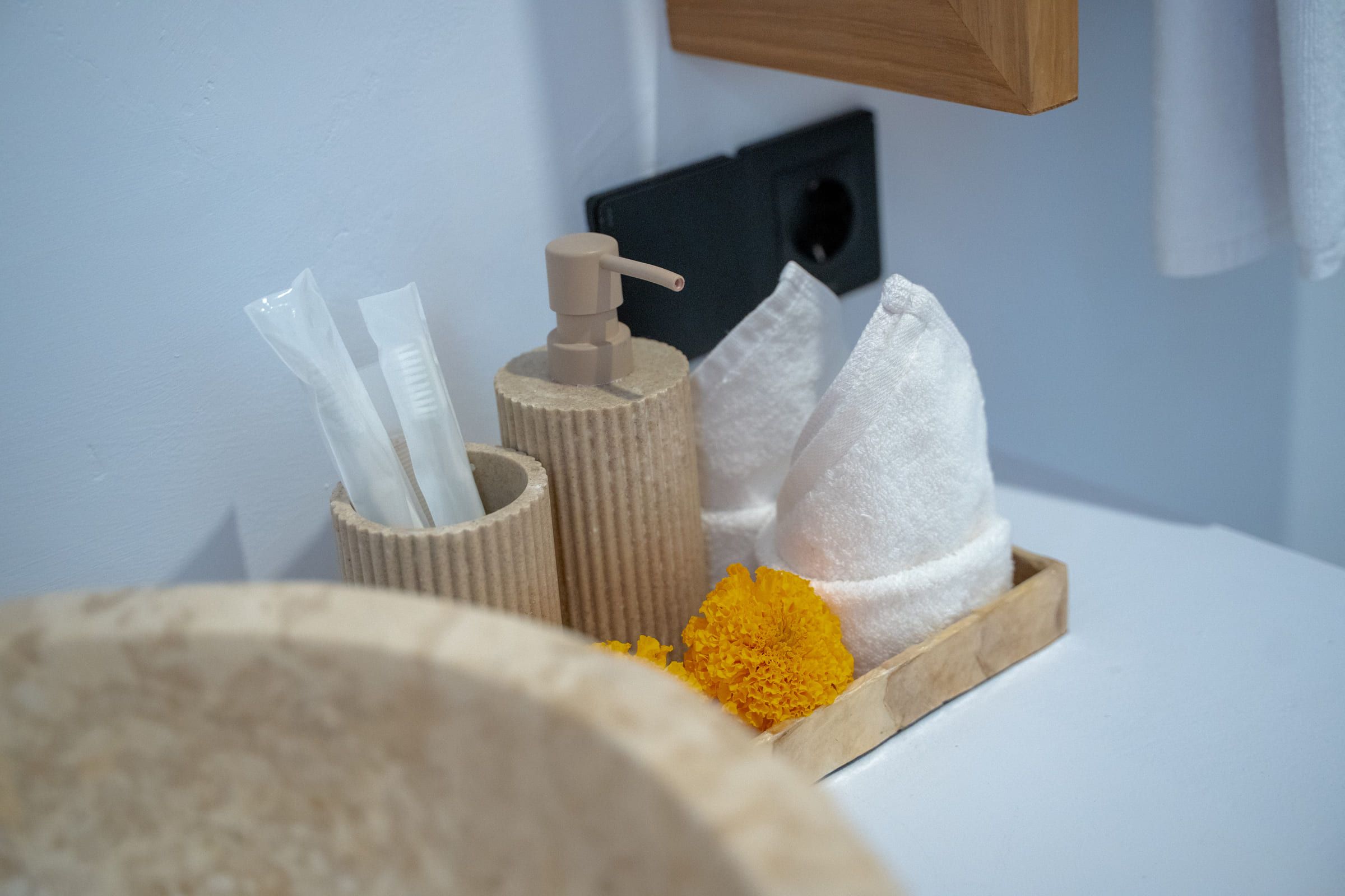 Bathroom amenities on a wooden tray: toothbrushes, a soap dispenser, folded towels and a marigold flower