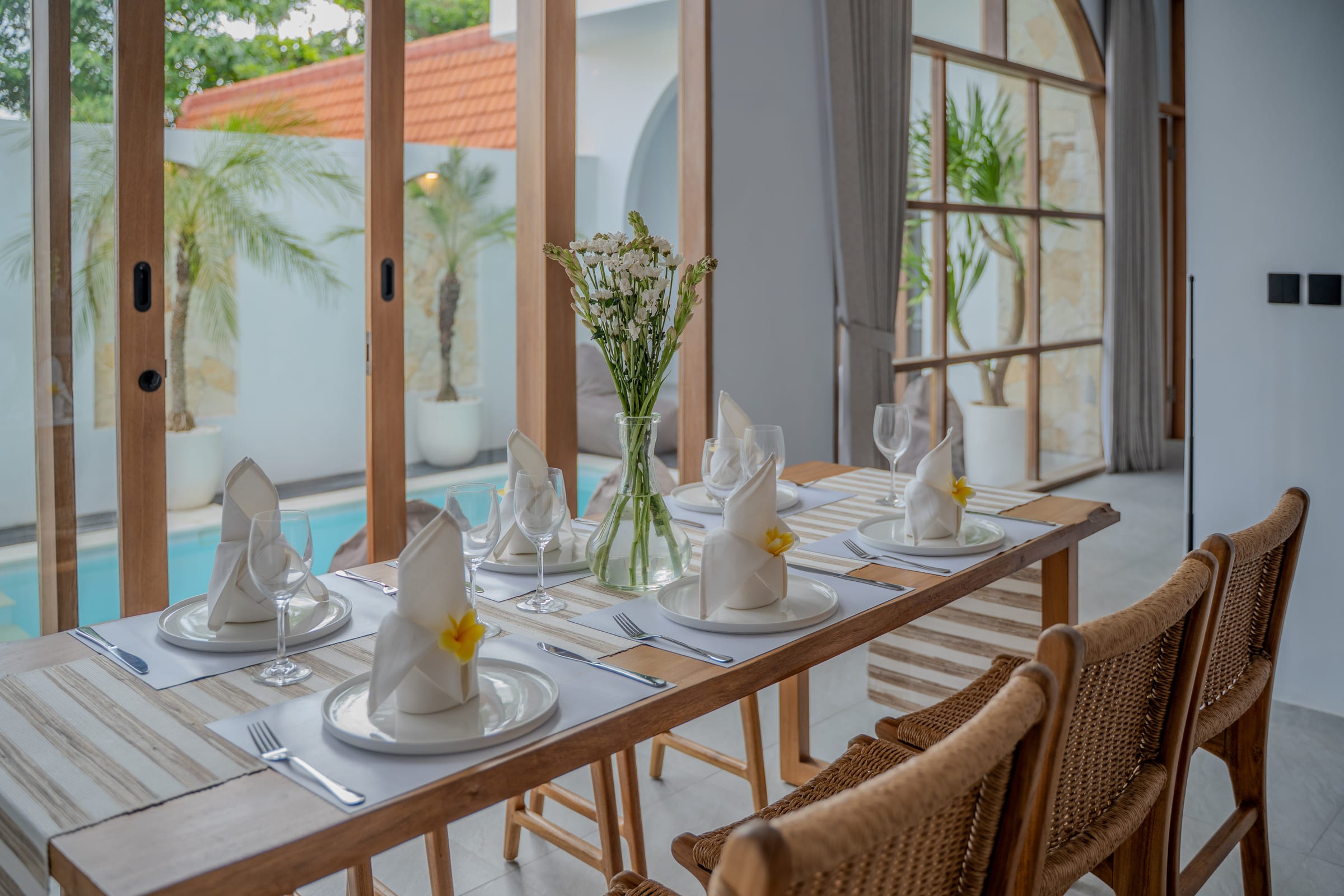 Dining table set with napkins and frangipani beside tall glass doors that look onto the pool