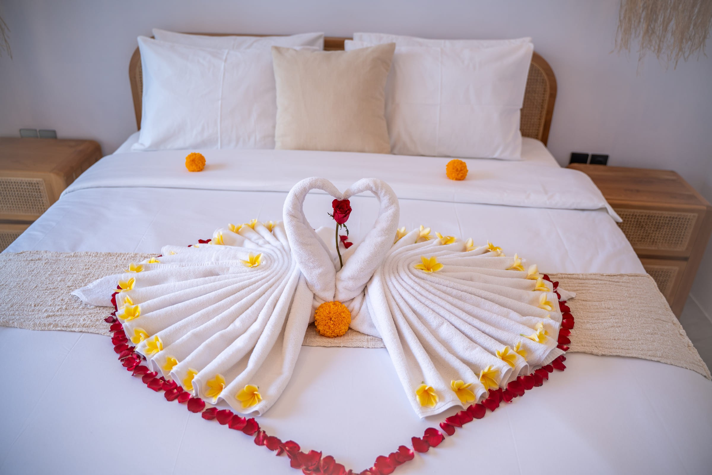Bed with white linens and a pair of folded towel swans forming a heart, decorated with frangipani and rose petals