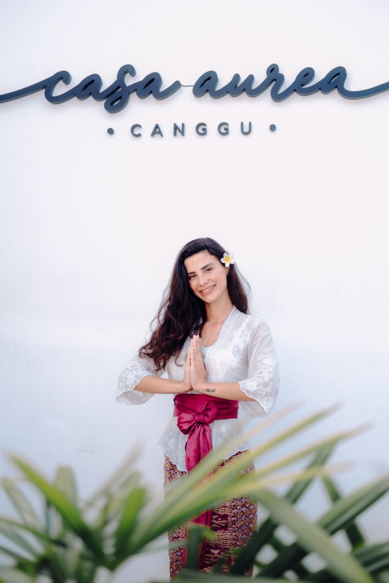 A smiling woman in traditional Balinese dress with a frangipani flower in her hair, greeting guests with folded hands beneath the Casa Aurea Canggu sign